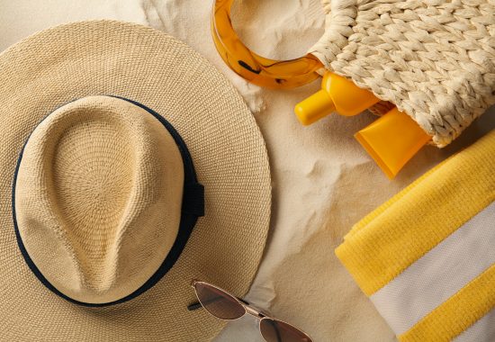 Straw hat, sunglasses, towel and bag with sunscreen on sand, flat lay