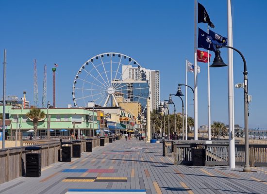 Along the wide scenic boardwalk of Myrtle Beach, with attractions, shops, and Ferris wheel in background.